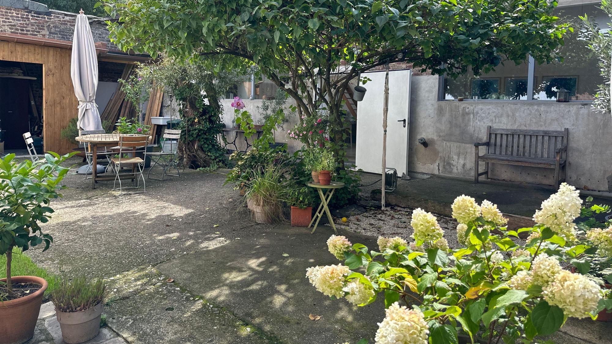Ein großer Innenhof mit verschiedenen Pflanztöpfen, einem Tisch mit Stühlen und einer Bank vor einer Wand sind zu sehen.