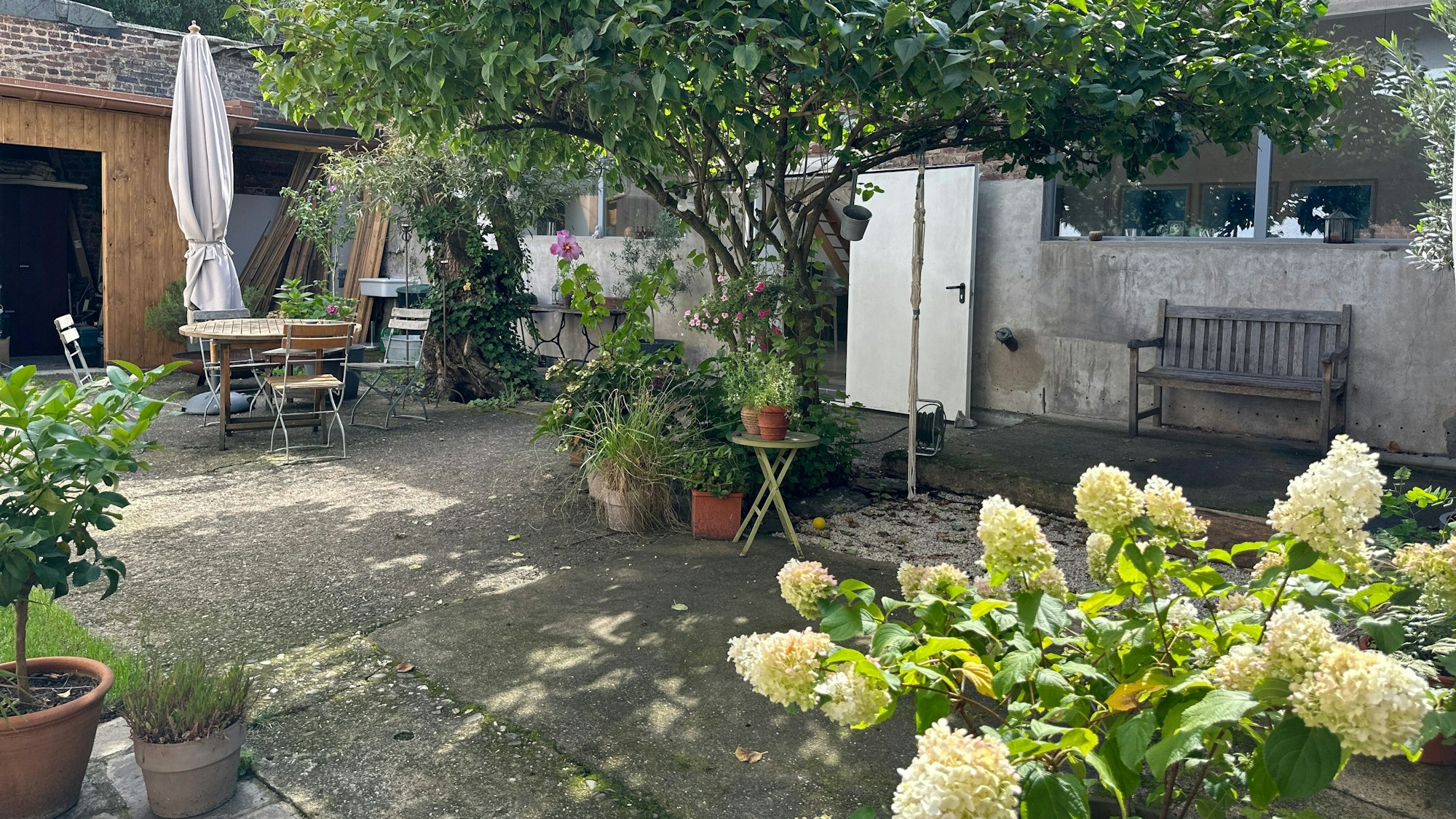 Ein großer Innenhof mit verschiedenen Pflanztöpfen, einem Tisch mit Stühlen und einer Bank vor einer Wand sind zu sehen.