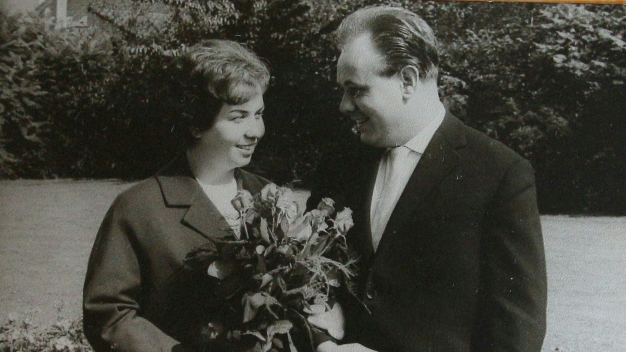 Eine Frau mit Blumenstrauß und ein Mann stehen nebeneinander und lächeln sich an.
