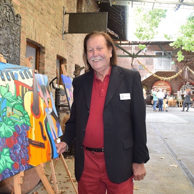 Ein Mann steht neben einer Staffelei, auf der ein buntes Bild steht.