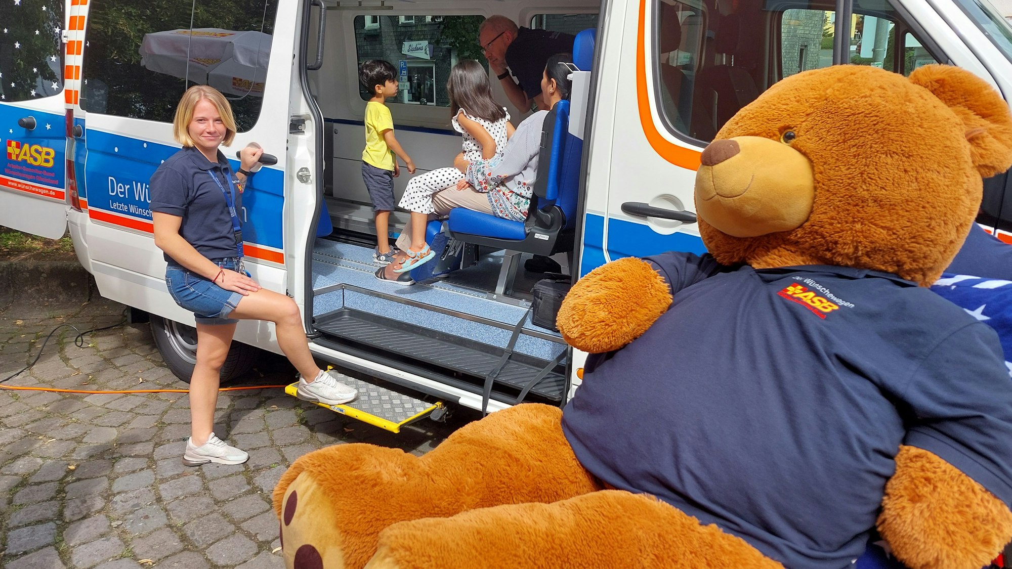 Ein weiß-blauer Transporter steht mit offener Schiebetür auf einem gepflasterten Platz, eine Frau hat den Fuß auf der Stufe, andere Menschen sitzen bgereits drin. Ein riesiger Plüschteddy im blauen ASB-Shirt sitzt davor.