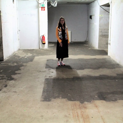 Eine Frau mit langen dunklen Haaren steht in einem nackten kellerartigen Raum.