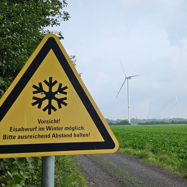 Ein Warnschild steht am Waldsaum neben einem Acker, im Hintergrund erheben sich drei Windräder.
