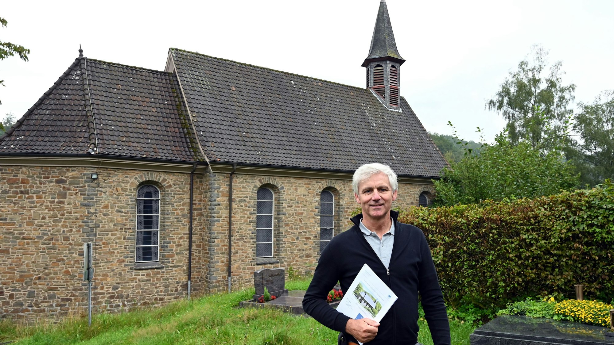 Der Makler Christoph Langer ist mit dem Verkauf der katholischen Kirche in der Morsbacher Ortschaft Ellingen beauftragt. Zum Grundstück gehört auch ein Teil des Friedhofs, auf dem der Reichshofer hier steht. In den Händen hält er das Exposé dieser ungewöhnlichen Immobilie.