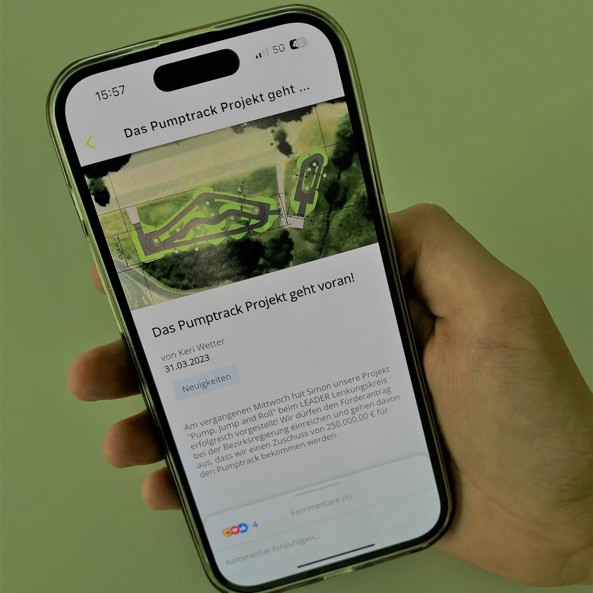 Eine Hand hält ein Smartphone. Auf dem Display steht: „Das Pumptrack-Projekt geht voran“.