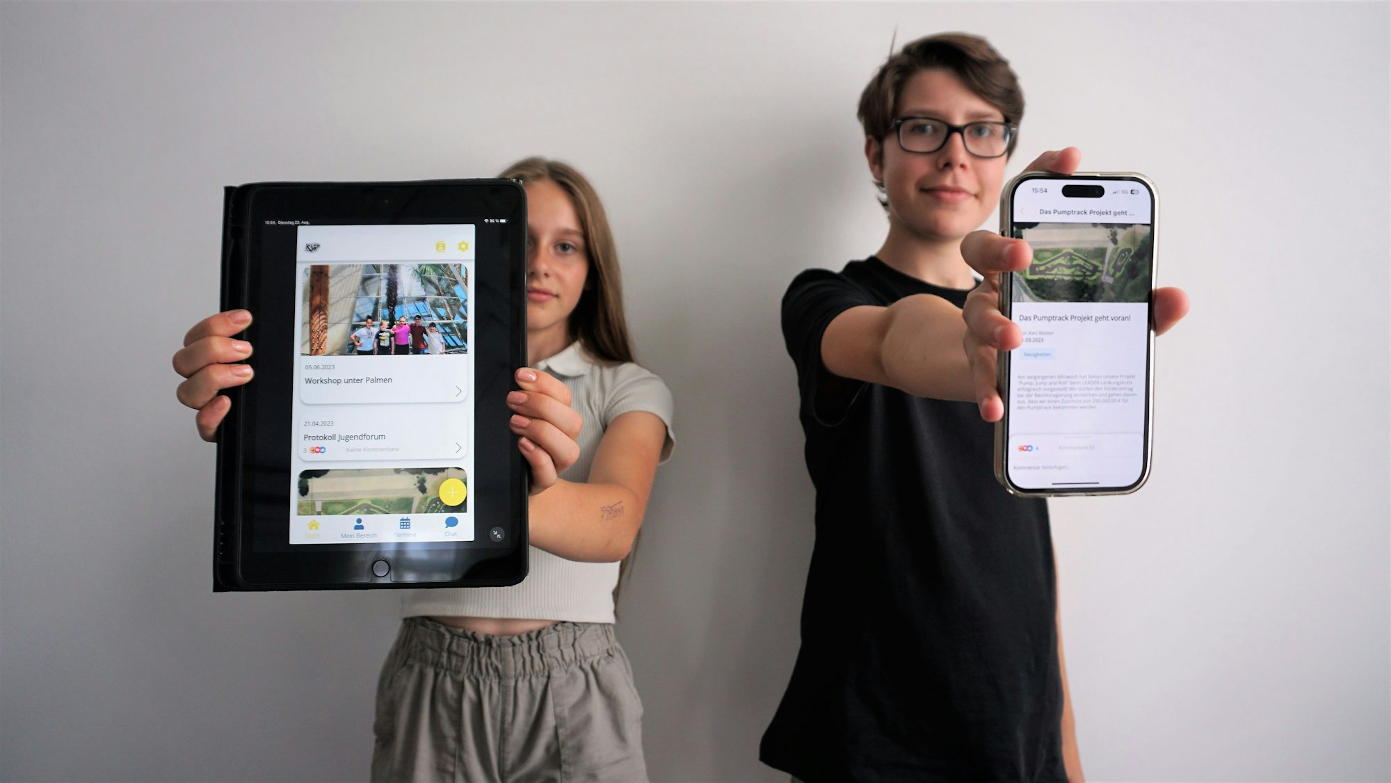 Sophie Schmidt und Simon Krämer halten mit ausgestreckten Armen ein Tablet und ein Smartphone in die Kamera