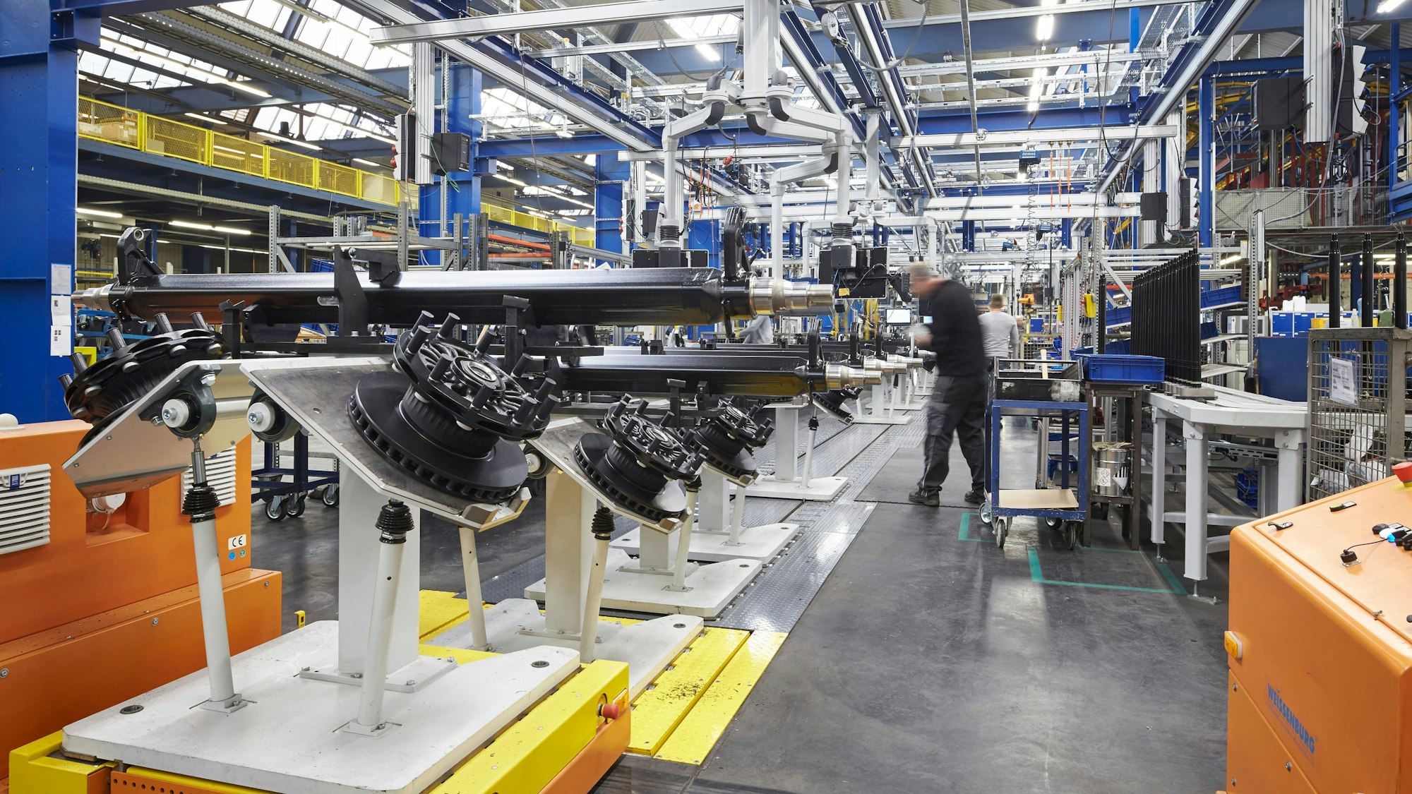 Man sieht Bestandteile einer LKW-Achse und einen Arbeiter in einer großen Fabrikhalle.