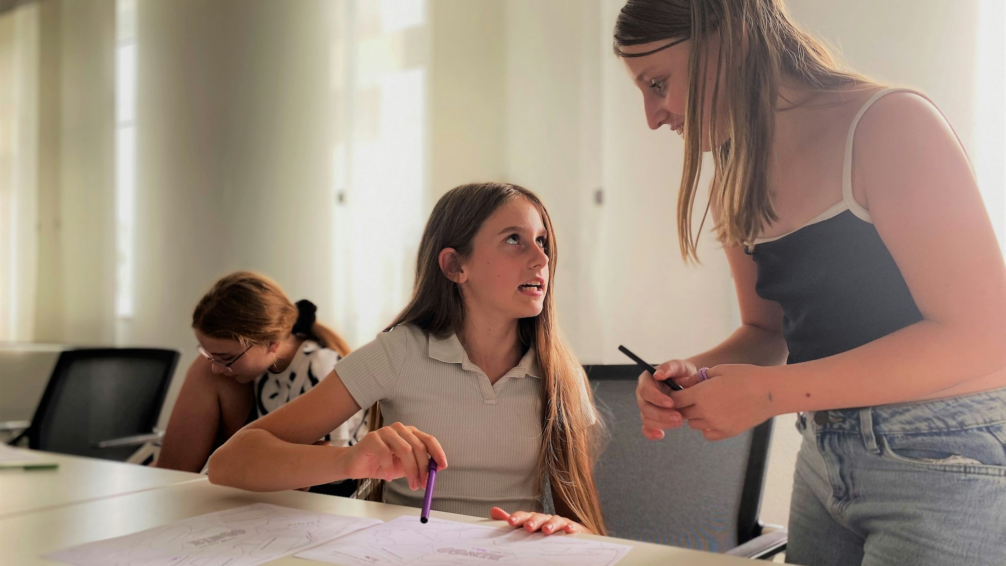 Zwei Mädchen diskutieren. Eines sitzt, eines steht. Beide haben einen Stift in der Hand.