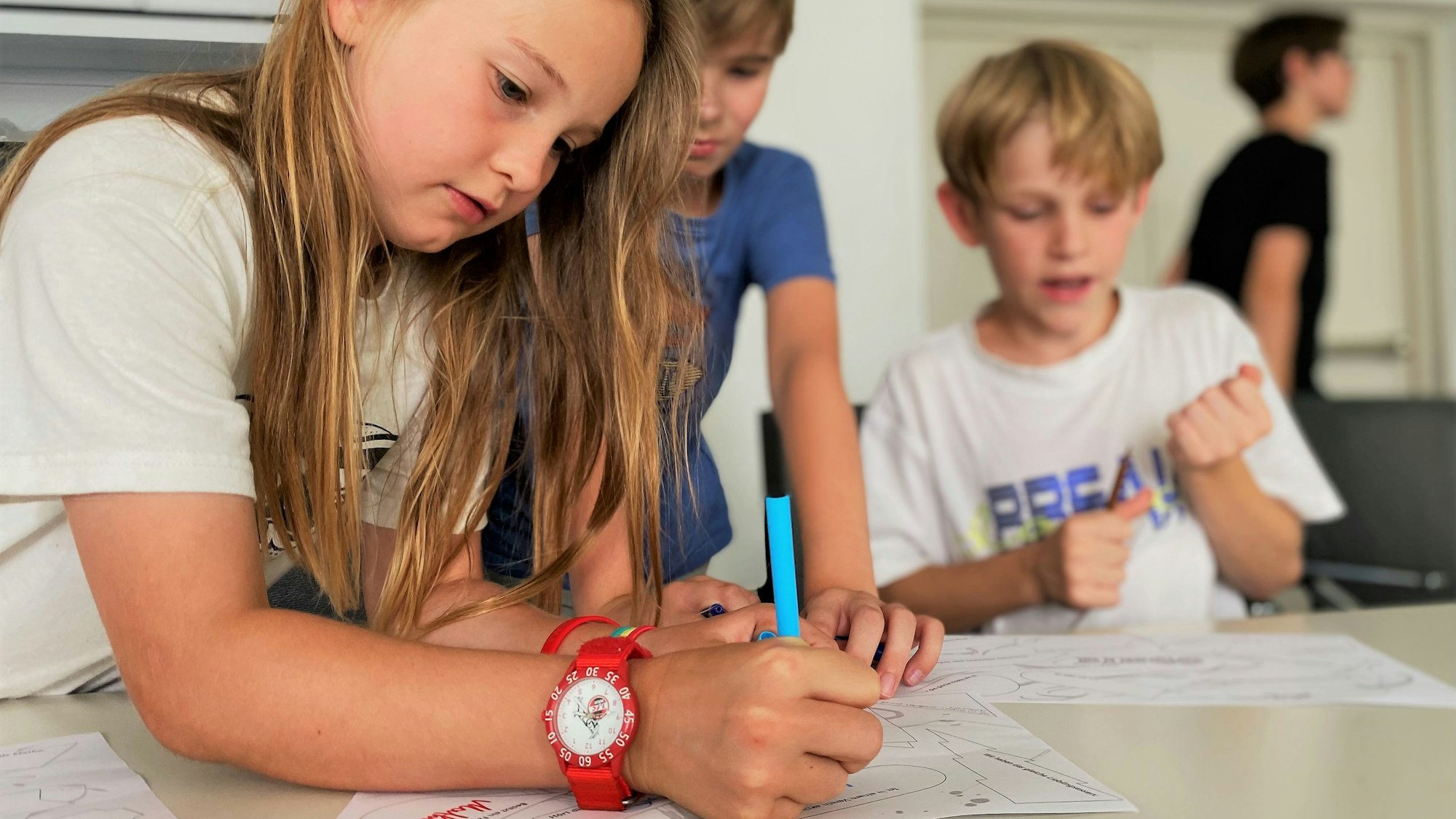 Ein Mädchen und zwei Jungs sind über Dokumente gebeugt. Das Mädchen schreibt.
