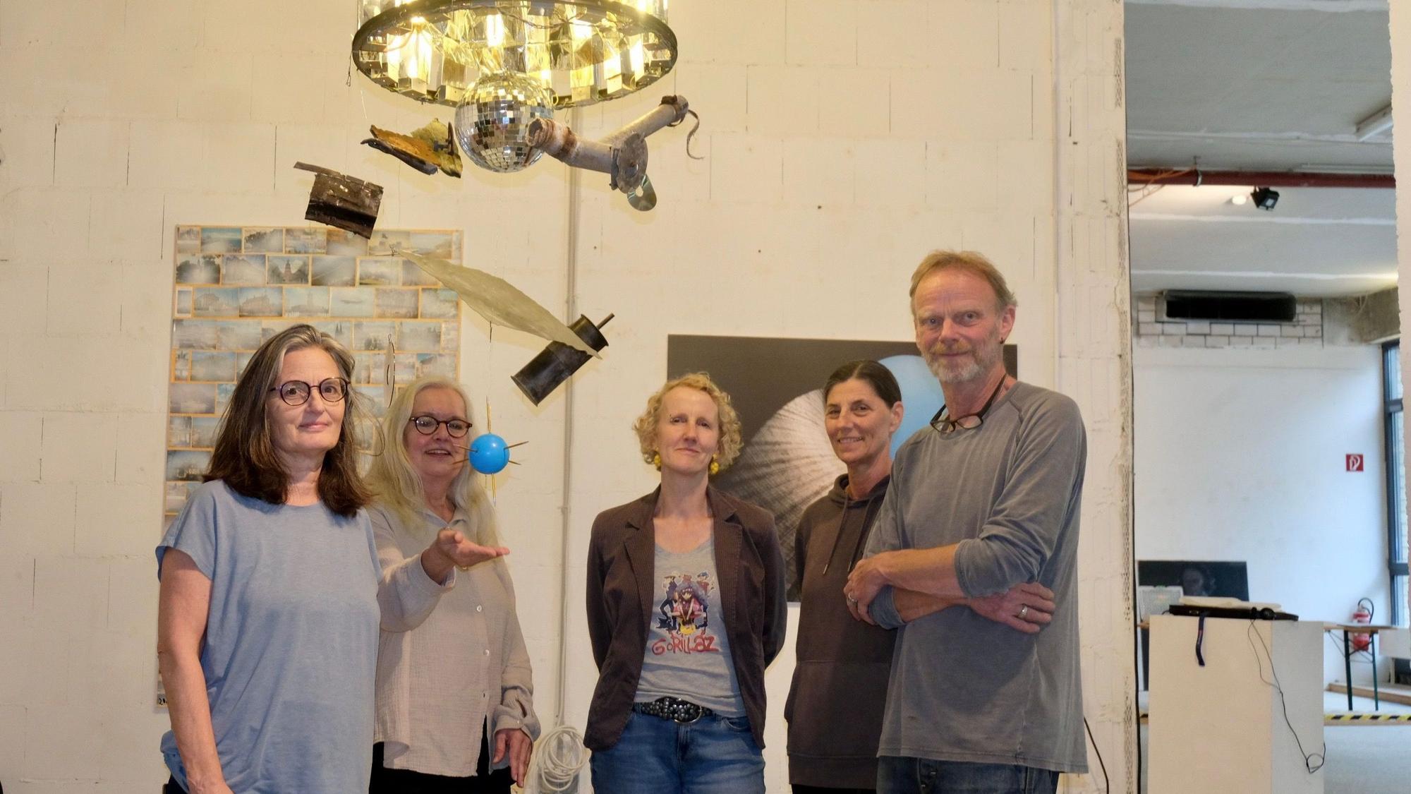 Vier Frauen und ein Mann stehen unter einem Kronleuchter, unter dem eine Diskokugel hängt, noch weiter unten baumeln eine blaue Kugel und verschiedene Objekte.