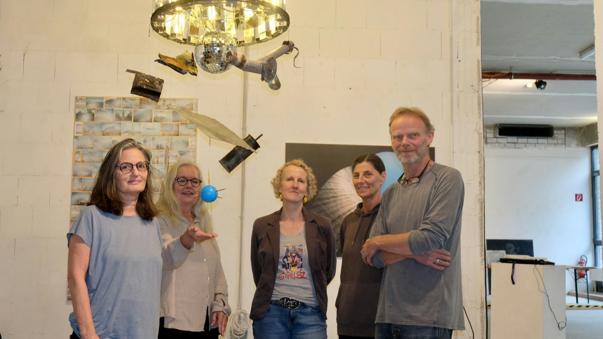 Vier Frauen und ein Mann stehen unter einem Kronleuchter, unter dem eine Diskokugel hängt, noch weiter unten baumeln eine blaue Kugel und verschiedene Objekte.