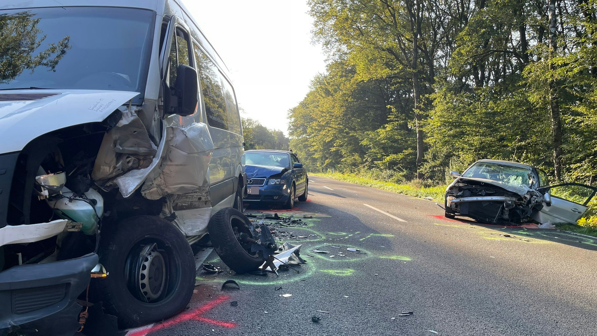 Die Fahrerseite eines Sprinters ist zerdrückt, das Rad abgerissen. Zwei weitere Fahrzeuge mit deutlichen Unfallspuren stehen weiter hinten auf der Straße, eines davon liegt halb im Graben. Markierungen sind auf der Straße aufgesprüht.