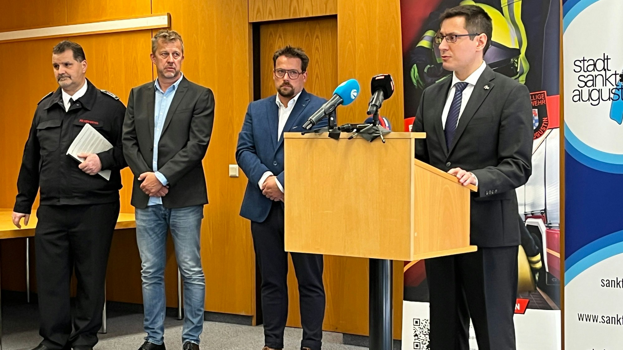 Vier Männer stehen in einem holzgetäfelten Raum, einer von ihnen an einem Rednerpult, auf dem mehrere Mikrofone stehen.