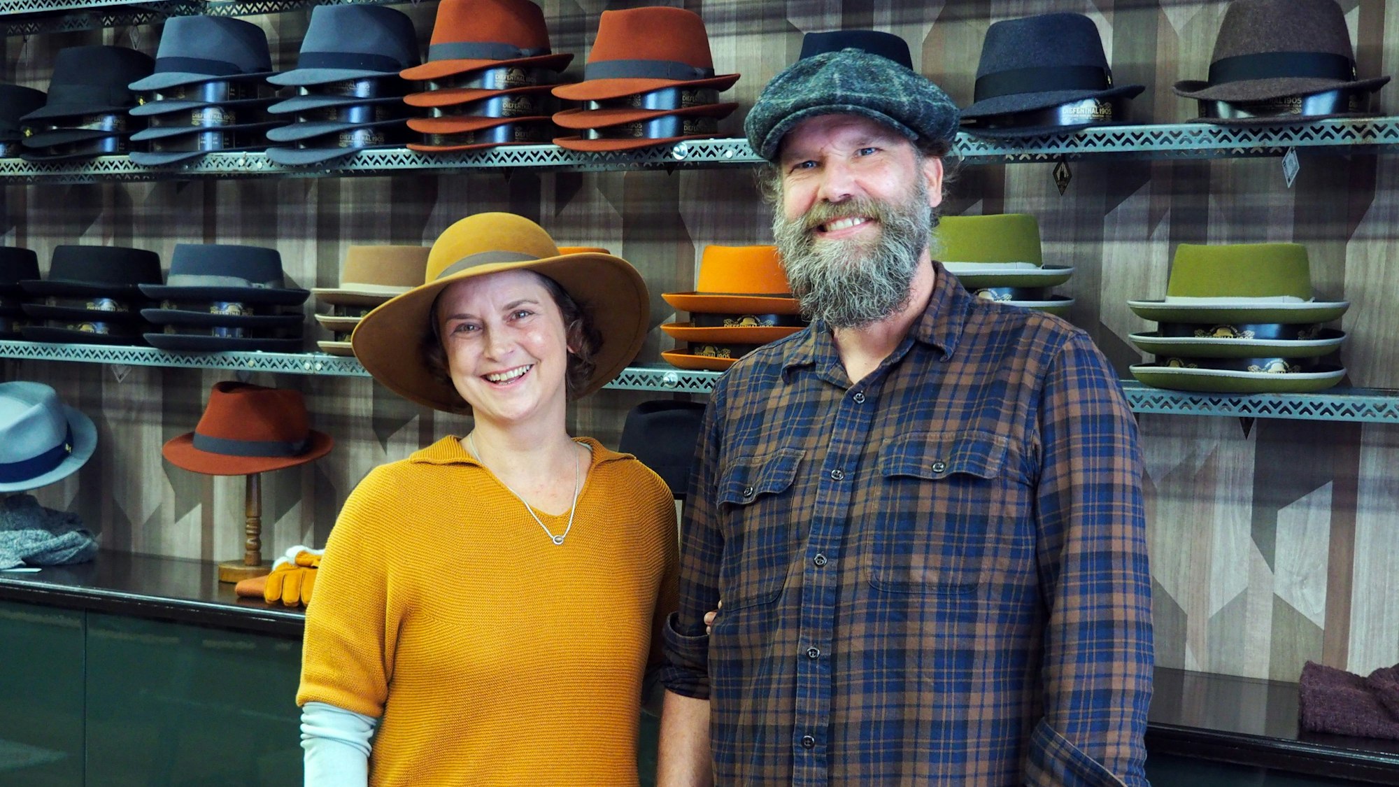 Eine Frau im gelben Pulli und ein Mann mit Bart und karierter Schiebermütze stehen vor einem Regal mit Hüten in vielen Farben.