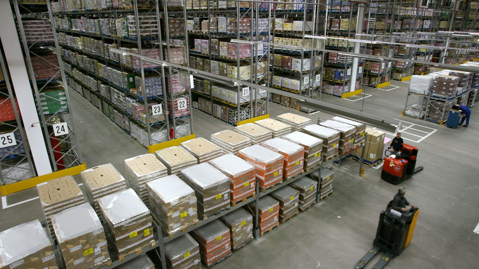 Zwei Gabelstapler fahren durch eine Halle, in der Regale mit Waren stehen.
