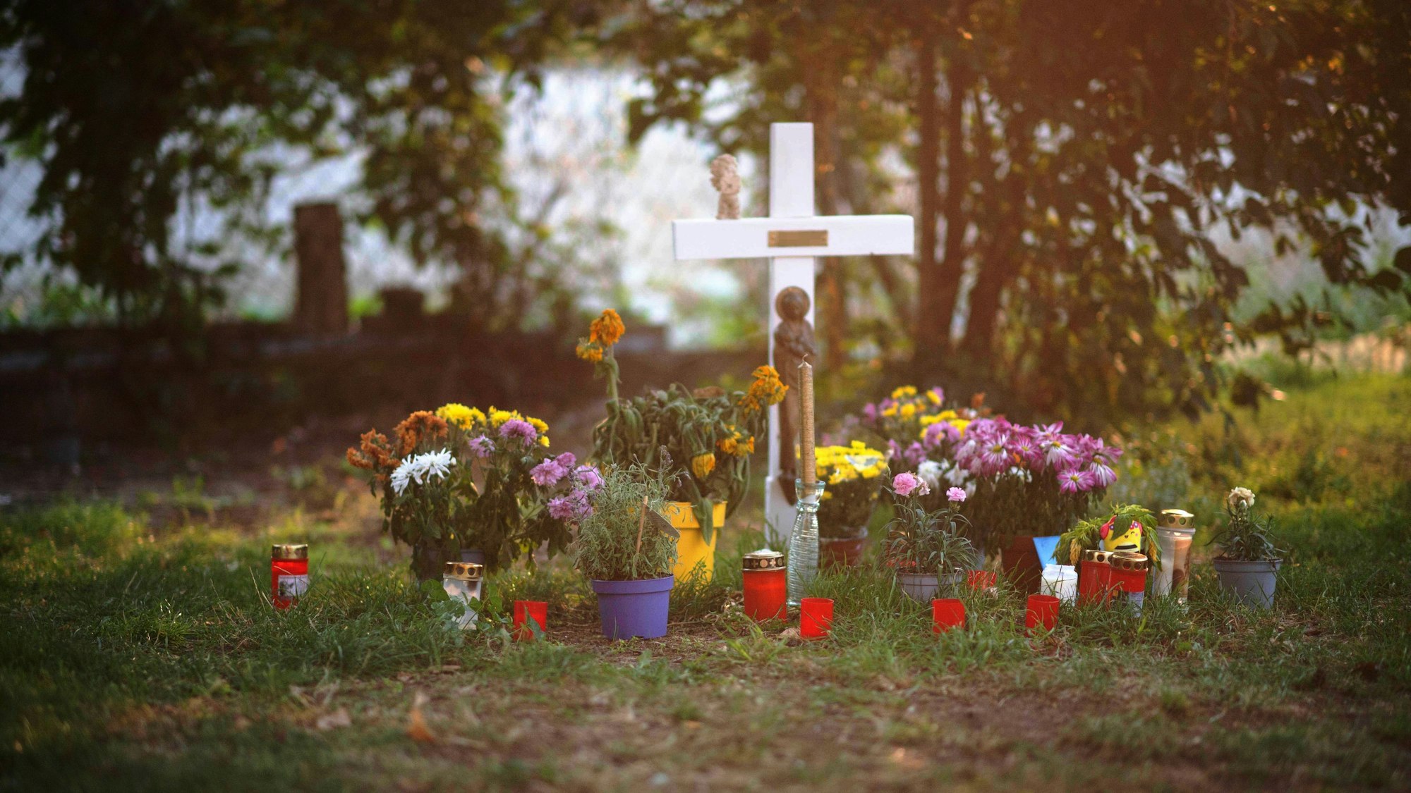 Das Bild zeigt ein weißes Holzkreuz, Blumen und Kerzen. Sie erinnern an den Tod eines Wohnungslosen.