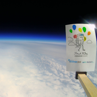 Die gekrümmte Erdoberfläche ist unter einer Wolkendecke vor dem dunklen Hintergrund des Weltalls zu sehen. Rechts ragt eine Karte mit der Aufschrift Paul-Klee-Gymnasium ins Bild.