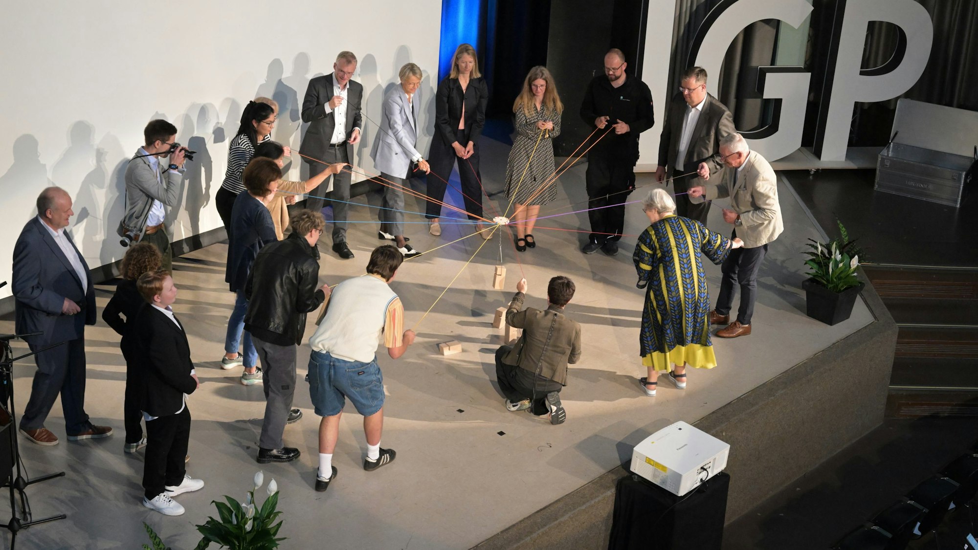 Die Gäste der Integrierten Gesamtschule Paffrath ziehen gemeinsam an Fäden. Auf der Bühne sind unter anderem NRW- Bildungsministerin Dorothee Feller , Bürgermeister Frank Stein, Landrath Stephan Santelmann und Schulleiterin Angelika Wollny