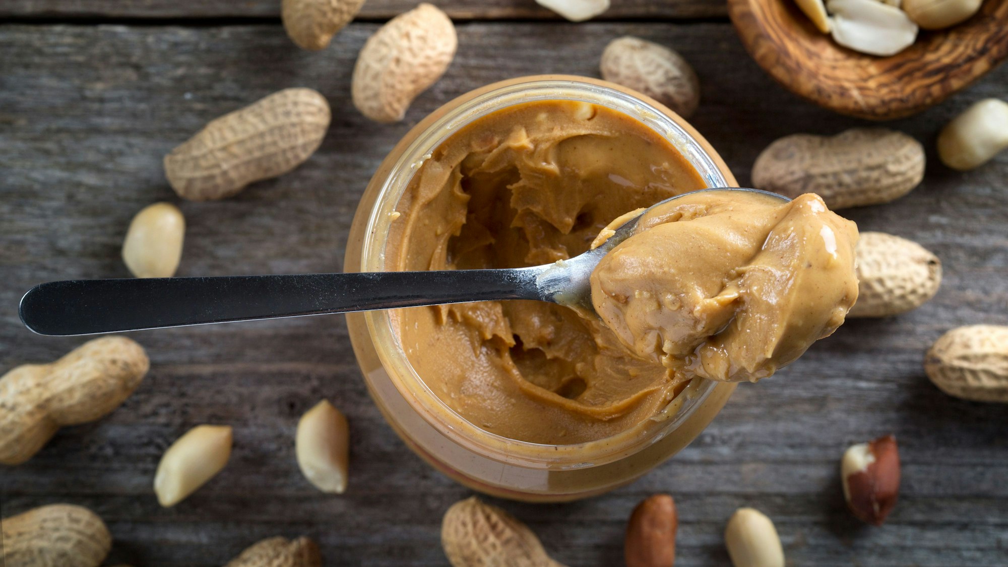Ein Löffel Erdnussbutter liegt auf einem Glas Erdnussbutter. Um das Glas herum liegen Erdnüsse auf einem hölzernem Untergrund.