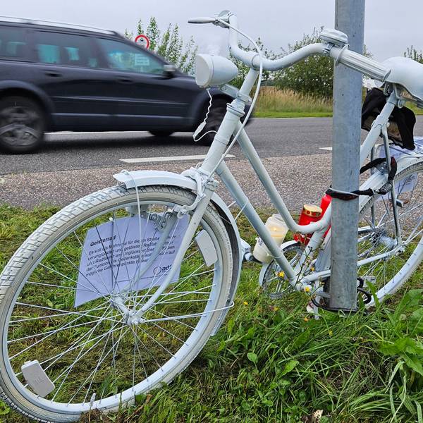 Ein komplett weiß angemaltes Fahrrad ist an einem Mast neben einer Straße angekettet, ein Auto fährt vorbei.
