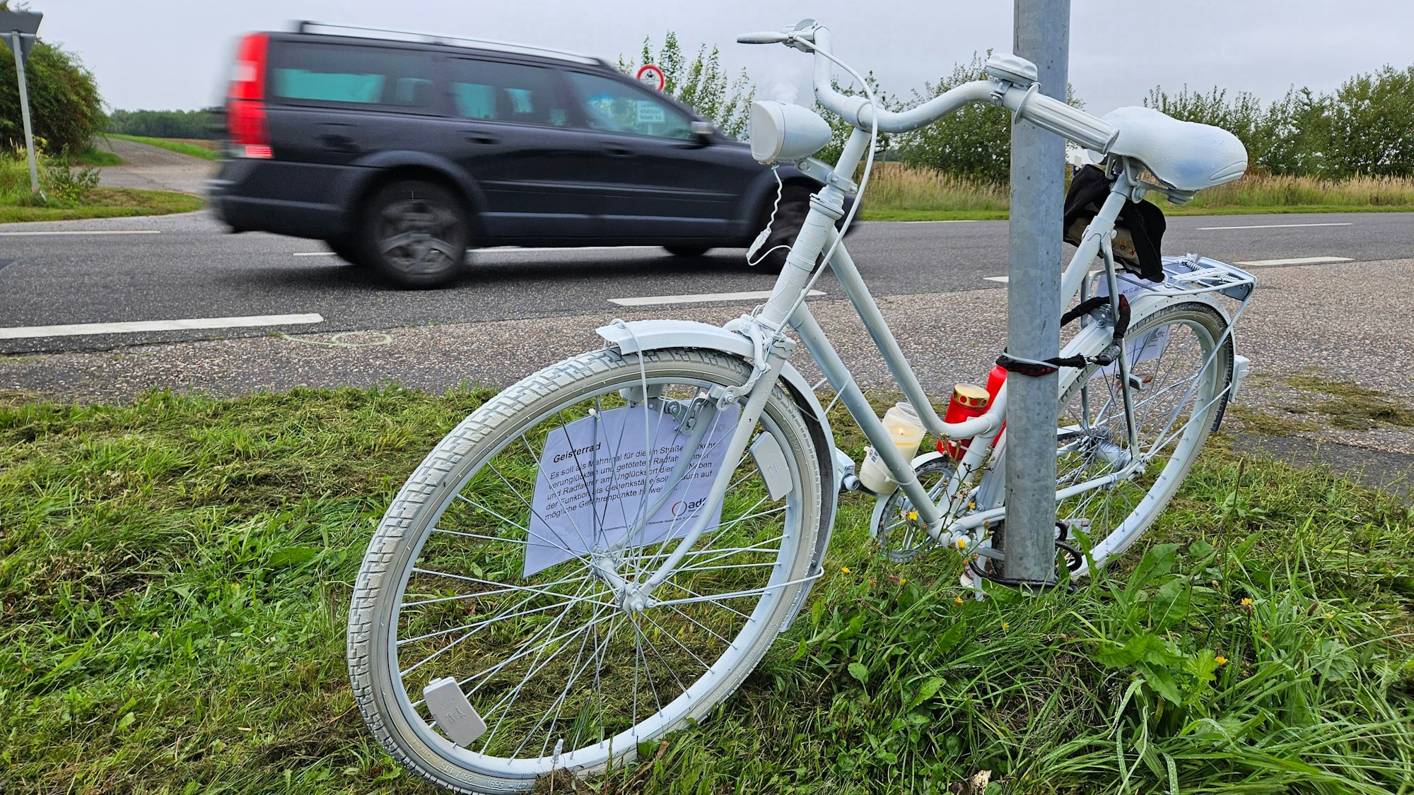 Ein komplett weiß angemaltes Fahrrad ist an einem Mast neben einer Straße angekettet, ein Auto fährt vorbei.