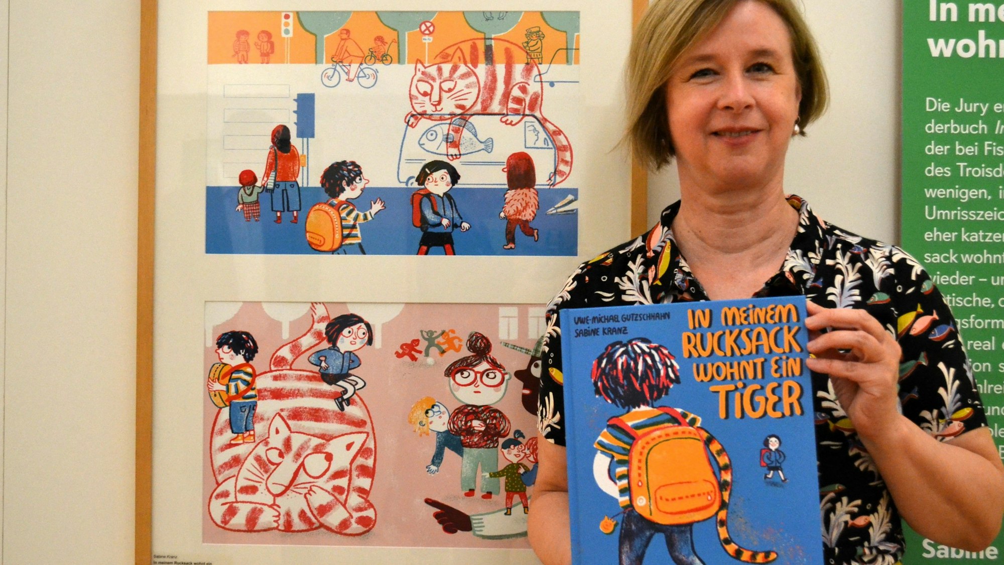 Eine blonde Frau hält das blaue Bilderbuch. Neben ihr hängen zwei Illustrationen aus ihrem Buch, die den katzenähnlichen Tiger mit Kindern zeigen.