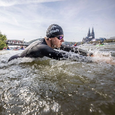 Ein Triathlet beim Schwimmen im Rhein, er trägt einen Neoprenanzug, eine Badekappe und eine Schwimmbrille. Der Kölner Dom und die Hohenzollernbrücke im Hintergrund.
