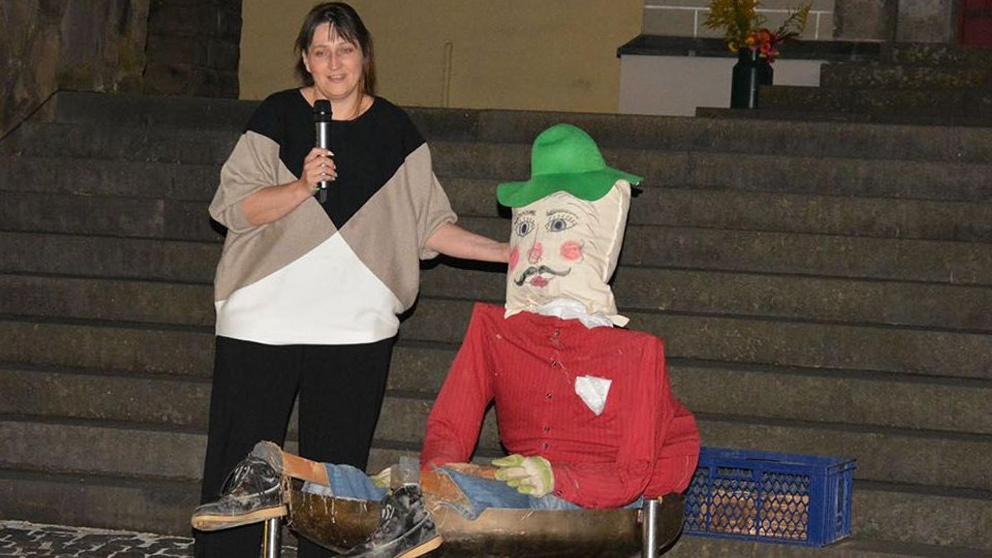 Eine Frau mit Mikrofon neben einer großen Stoffpuppe, die einen grünen Hut trägt.