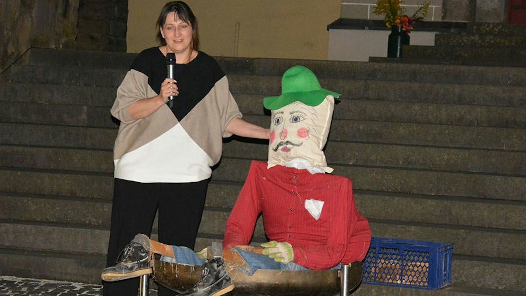 Eine Frau mit Mikrofon neben einer großen Stoffpuppe, die einen grünen Hut trägt.