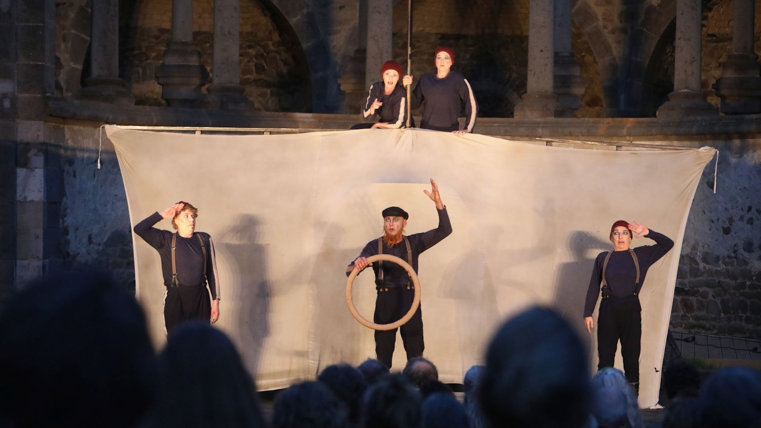 Drei Schauspieler vor einem Segel, zwei obenauf — im Hintergrund ist die Ruine des Abteichors von Kloster Heisterbach zu erkennen.