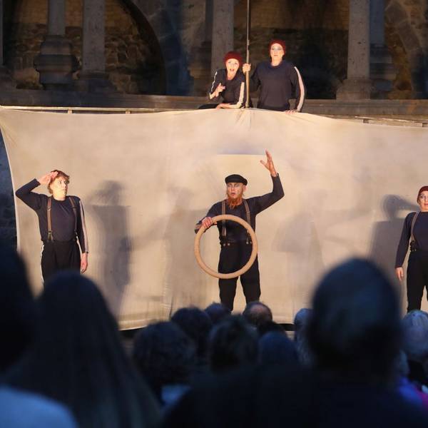 Drei Schauspieler vor einem Segel, zwei obenauf — im Hintergrund ist die Ruine des Abteichors von Kloster Heisterbach zu erkennen.