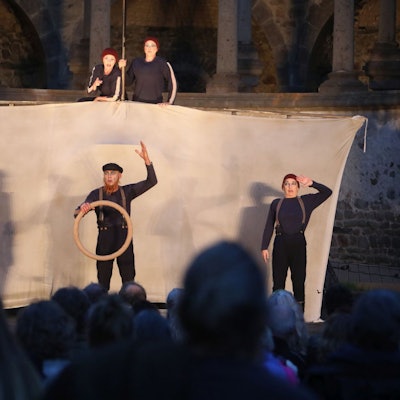 Drei Schauspieler vor einem Segel, zwei obenauf — im Hintergrund ist die Ruine des Abteichors von Kloster Heisterbach zu erkennen.