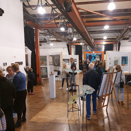 Eine große Ausstellungshalle mit Holzboden und einem Dach aus Stahlträgern. Skulpturen und Staffeleien stehen im Raum, an den Wänden hängen Bilder. Viele Menschen betrachten die Kunstwerke.