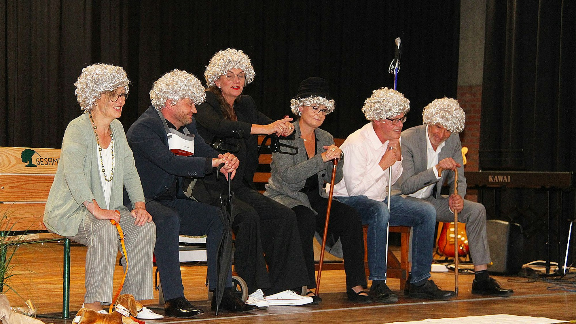Sechs Lehrerinnen und Lehrer sitzen mit Perücken auf einer Bank.