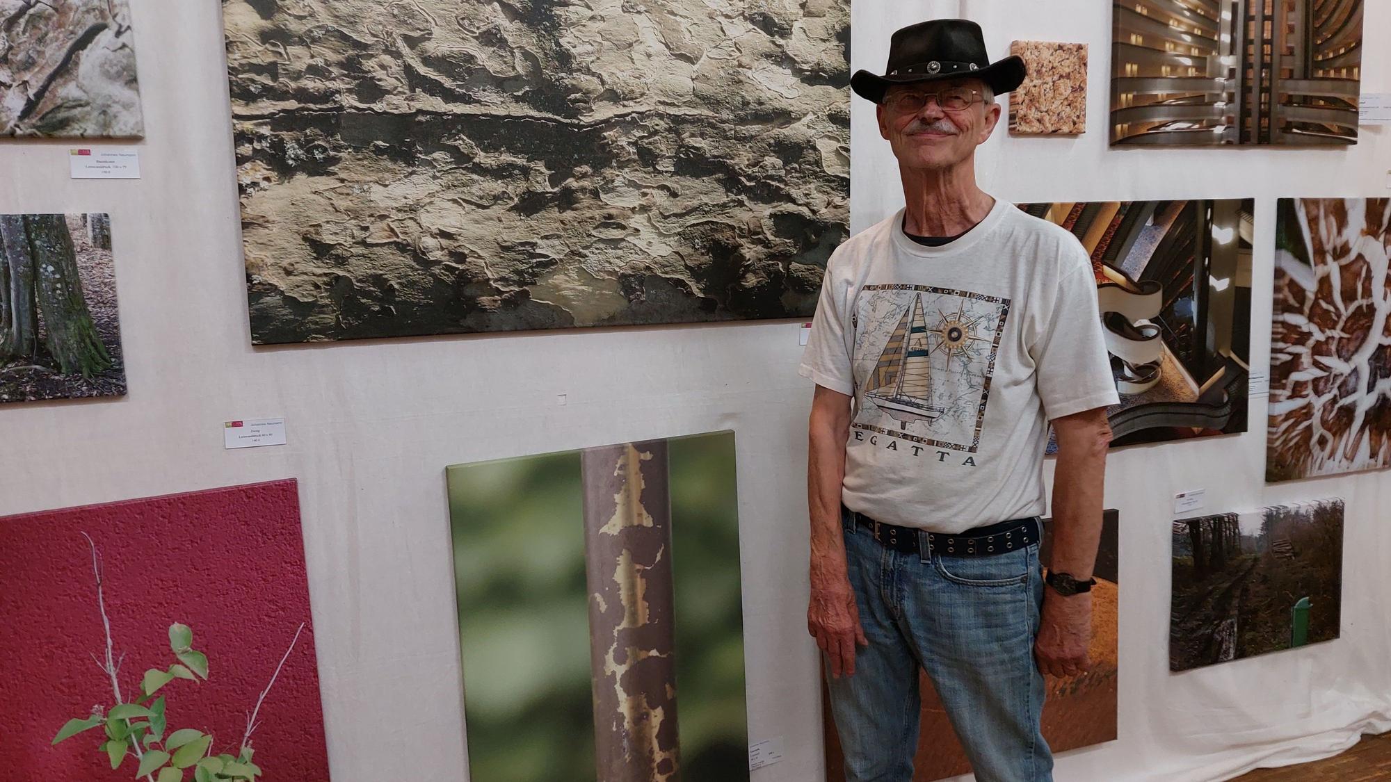 Ein älterer Mann mit Cowboyhut vor großen Fotoggrafien, die Details von Wänden, Pflanzen oder Rohren zeigen.