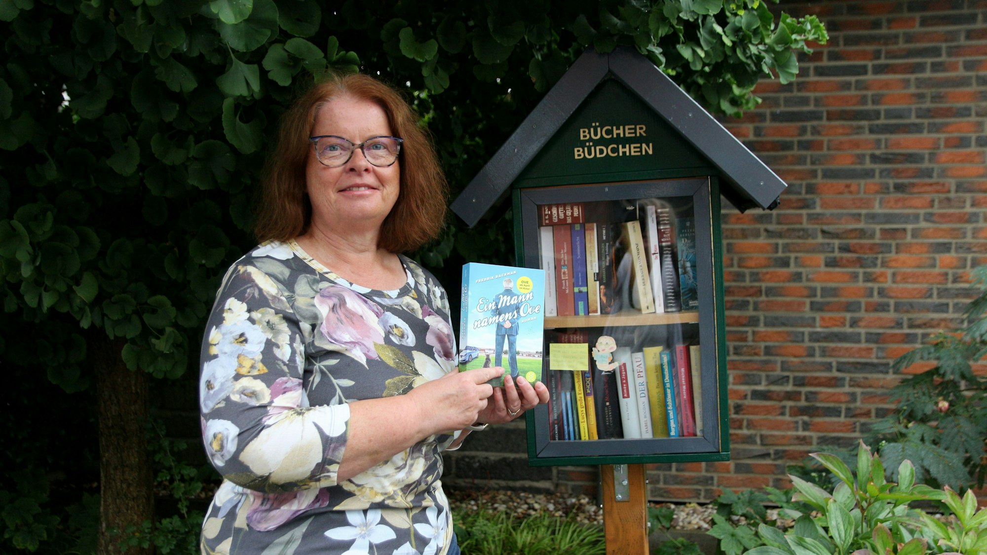 Eine Frau steht mit einem Buch in der Hand vor einem kleinen Bücher-Schrank.