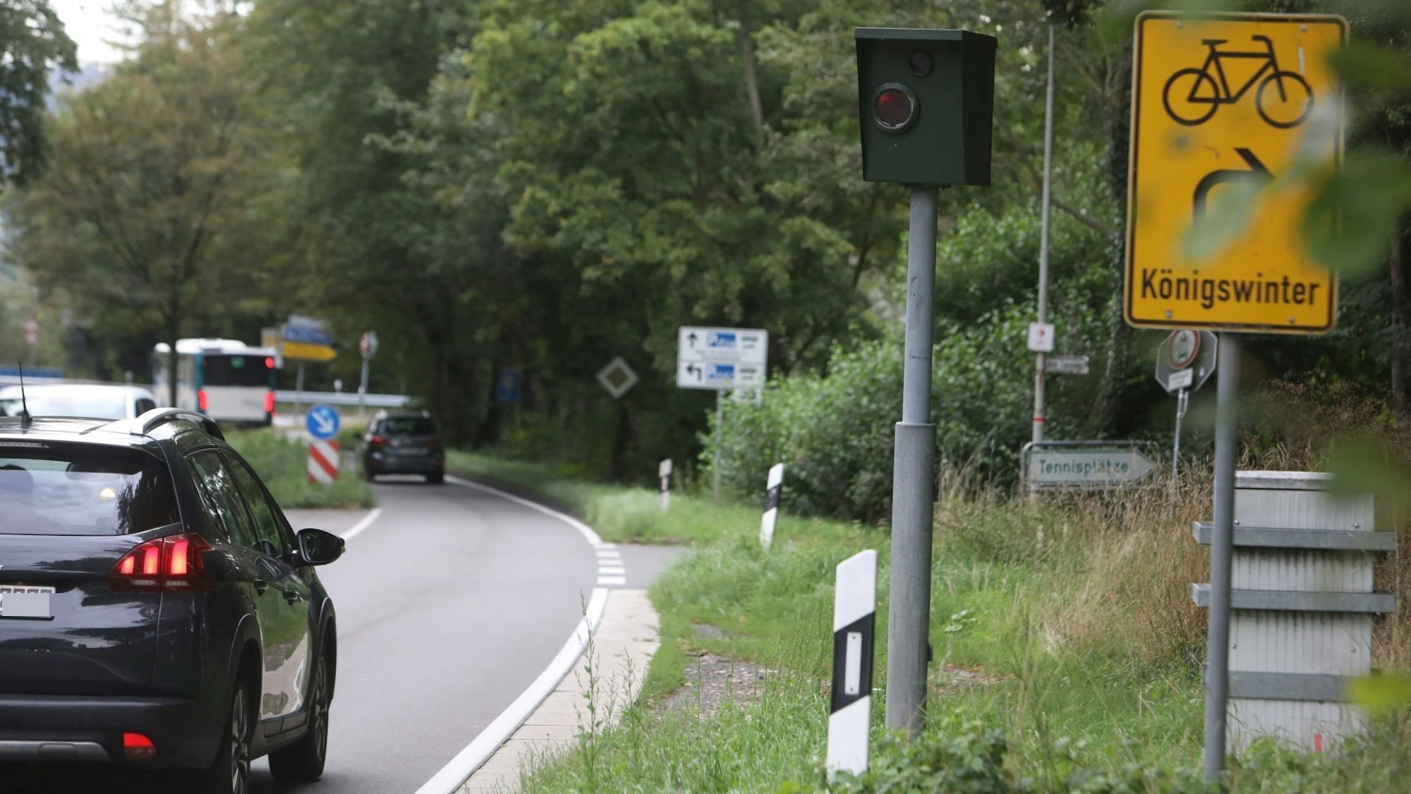 Eine gebogene Straße, auf der ein Auto fährt. Am Straßenrand steht ein grüner Radarkasten, daneben ein gelbes Schild mit der Richtung Königswinter.