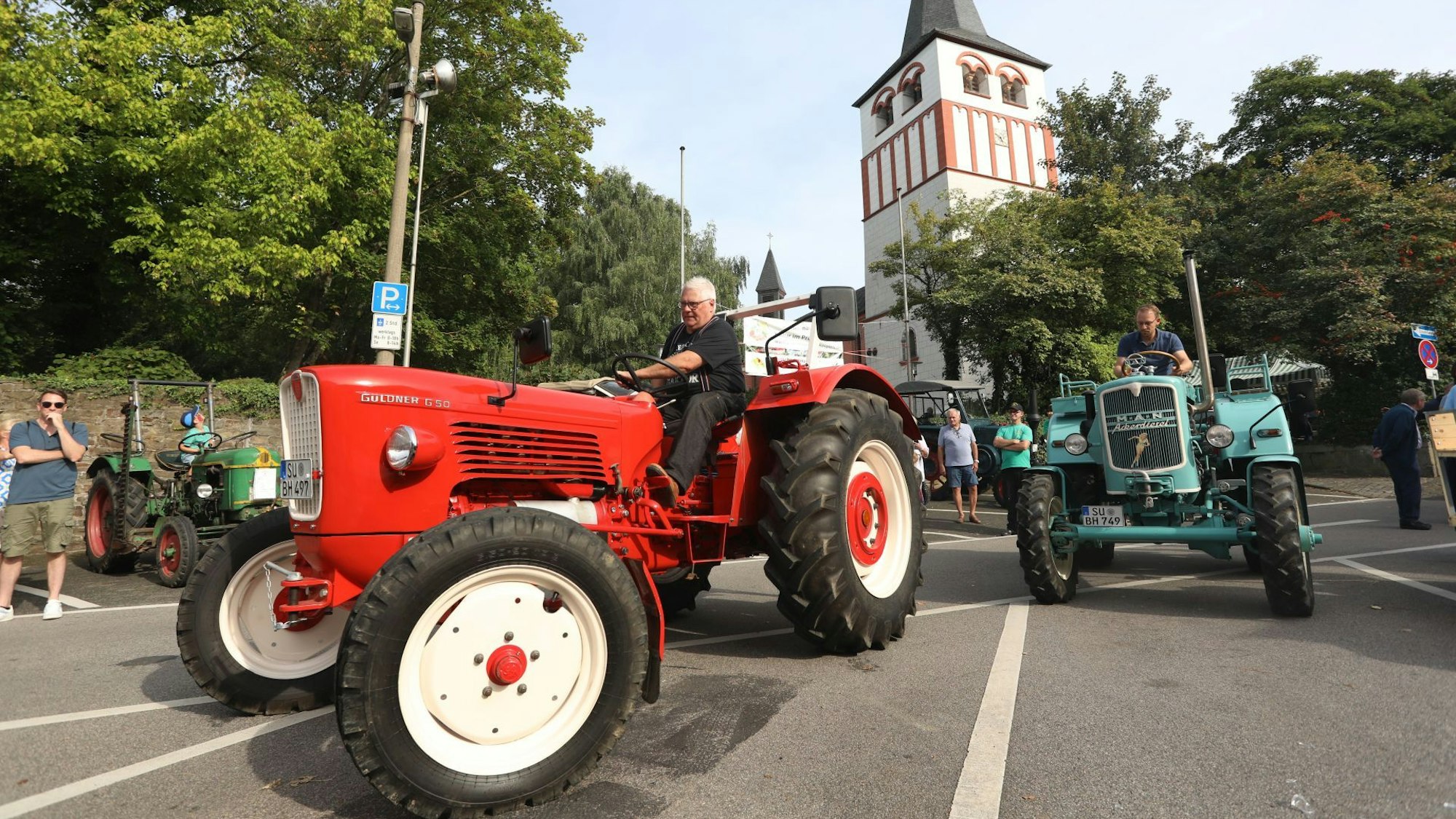Zwei Oldtimer-Trecker, ein roter und ein blauer, fahren über die Straße. Im Hintergrund ist die Kirche von Oberpleis.