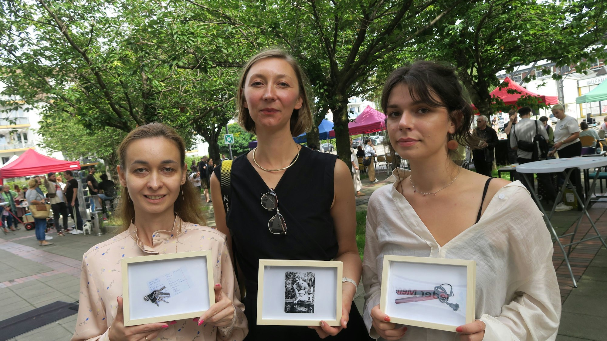Drei Frauen halten kleinformatige Bilder in der Kamera, hinter ihnen ist ein baumbestandener Platz mit vielen Menschen.