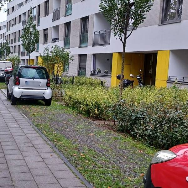 Vor einem Wohnblock parken Autos auf einer Schotterfläche neben Rad- und Fußweg.