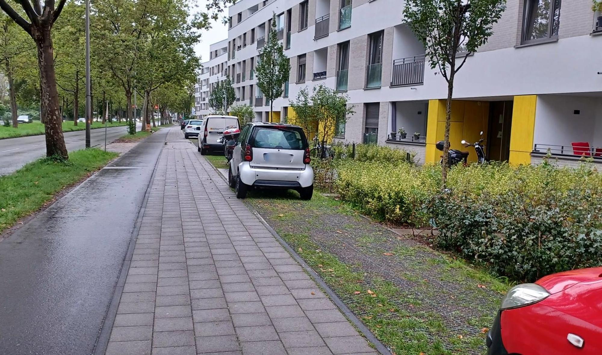 Vor einem Wohnblock parken Autos auf einer Schotterfläche neben Rad- und Fußweg.