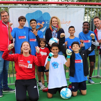 Junge Fußballer von dem Kölner Verein „Kidsmiling“ und Vereinsmitarbeiterinnen und -Mitarbeiter stehen vor einem Tor in Siegerposen.
