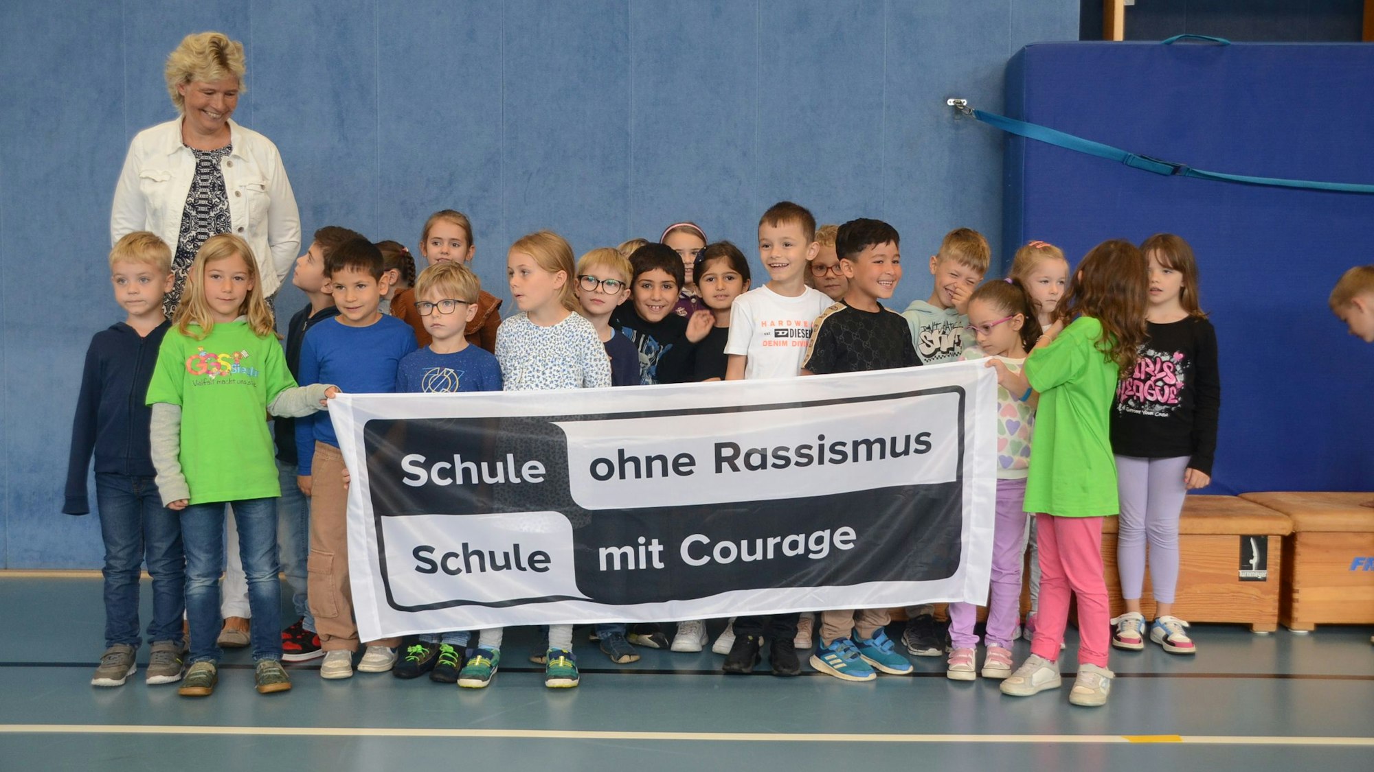 Jungen und Mädchen aus einer Grundschulklasse stehen vor einer blauen Wand in einer Turnhalle. Sie halten ein Transparent in Schwarz und Weiß hoch. „Schule mit Courage - Schule ohne Rassismus“ steht darauf. Eine blonde Fraue mit heller Jacke schaut auf die Kinder, sie lächelt.