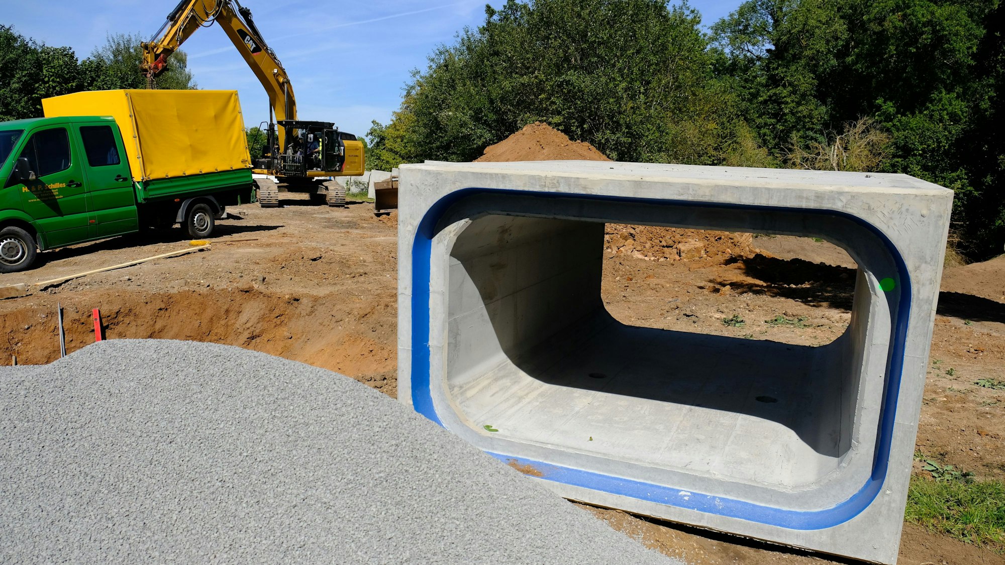 Ein kastenförmiges Rohr liegt auf einer Baustelle in Satzvey. Ein Haufen Splitt sowie im Hintergrund ein Lkw und ein Bagger sind zu sehen.