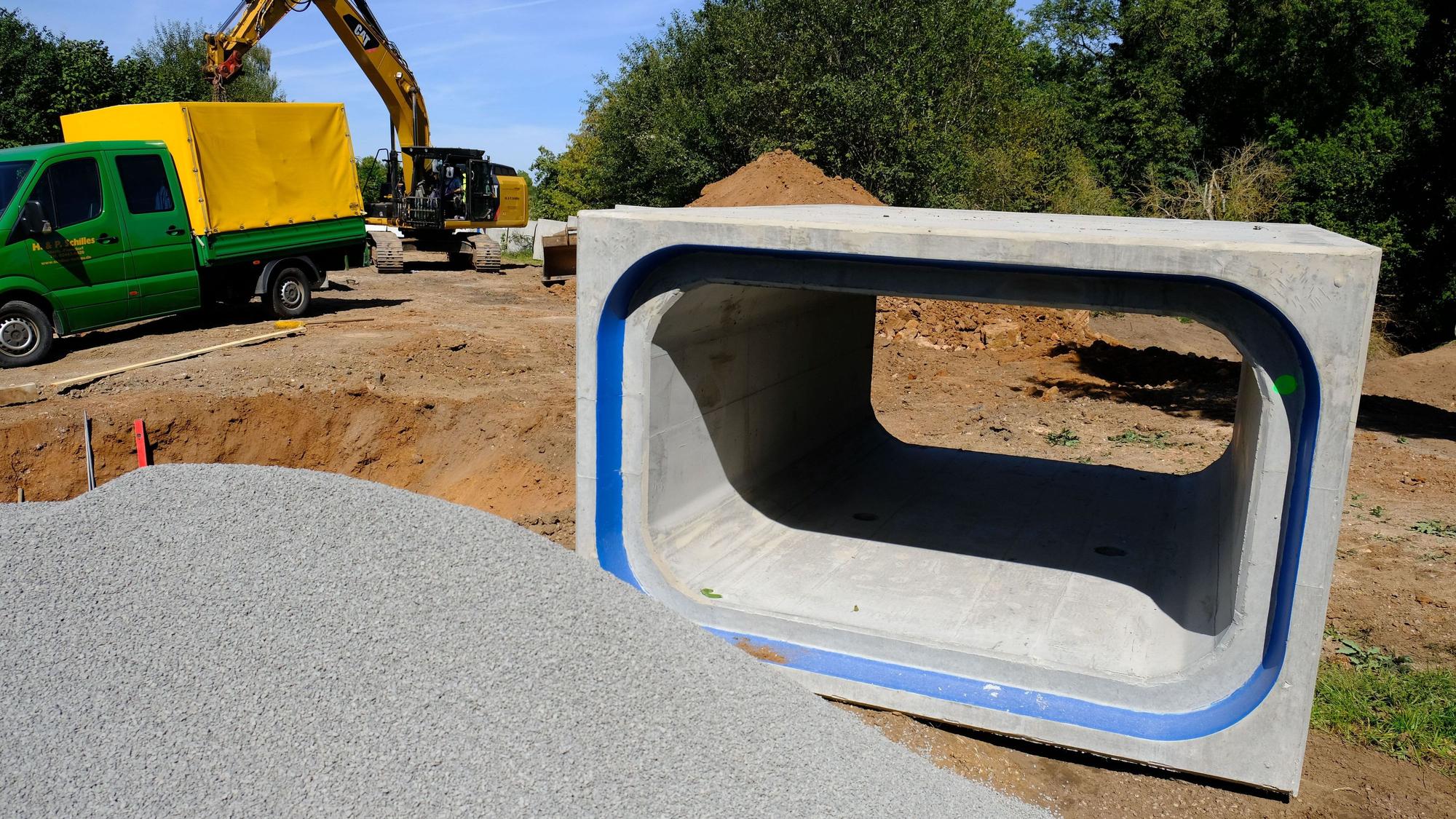An einer Baustelle in Satzvey liegt ein kastenförmiges Betonrohr, das zum Hochwasserschutz eingesetzt wird. Im Hintergrund sind ein Bagger und ein Lkw zu sehen.