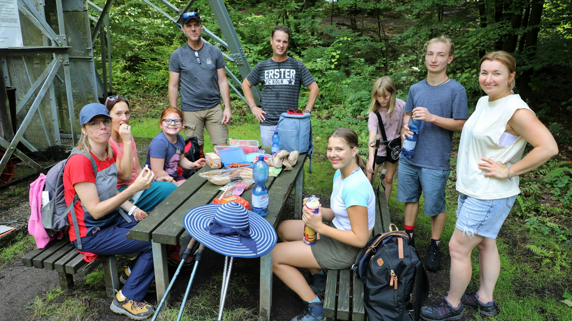 Drei Familien sitzen und stehen um einen Tisch unter freiem Himmel und machen Picknick.