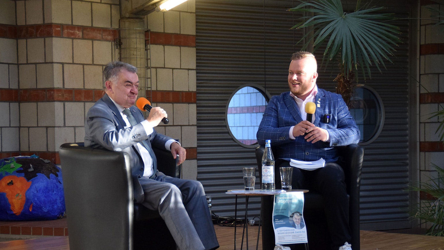 Herbert Reul im Gespräch und Dominik Pinsdorf sitzen auf einem Stuhl und halten ein Mikrofon in der Hand.