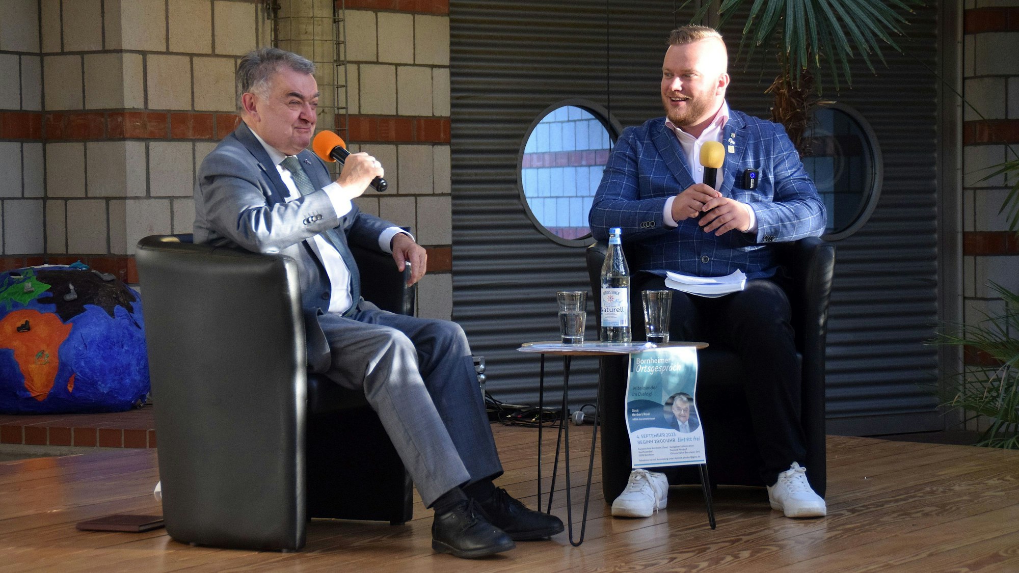 Herbert Reul im Gespräch und Dominik Pinsdorf sitzen auf einem Stuhl und halten ein Mikrofon in der Hand.