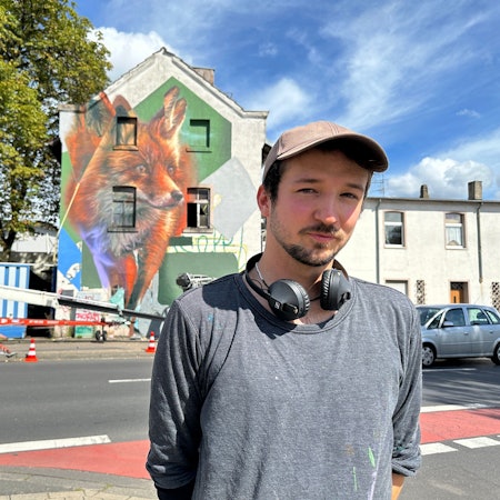 Ein junger Mann mit Käppi und Kopfhörern um den Hals steht an einer Straße. Im Hintergrund ist ein Gebäude zu sehen, auf dem ein großer Fuchs prangt.