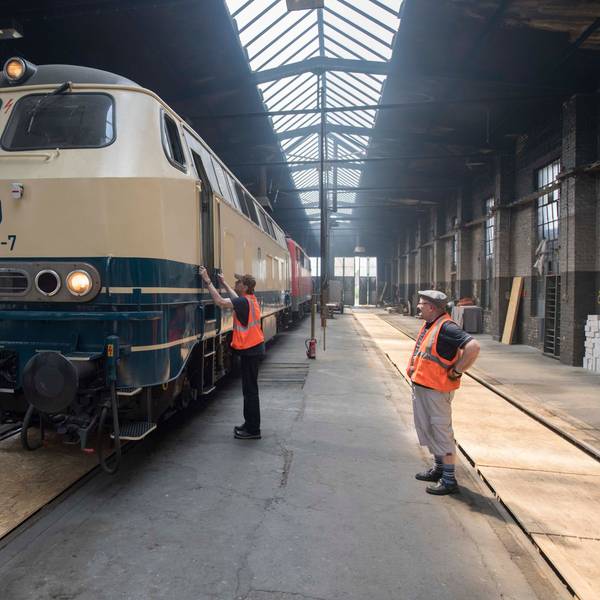 Zwei Männer in orangen Warnwesten stehen in einer großen Halle. Der Linke steht an einer historischen Lokomotive.